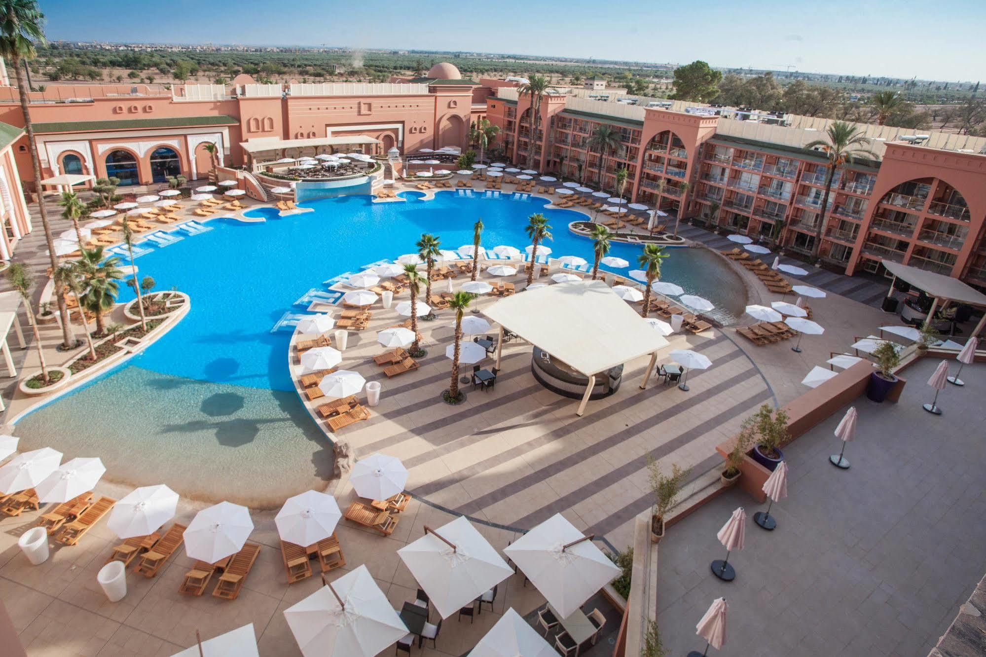 Savoy Le Grand Hotel Marrakech Μαρακές Εξωτερικό φωτογραφία The photo shows a large, luxurious hotel resort with a spacious swimming pool at its center. The pool area features several lounge chairs and umbrellas arranged around the water, providing a relaxing atmosphere for guests. Surrounding the pool are la